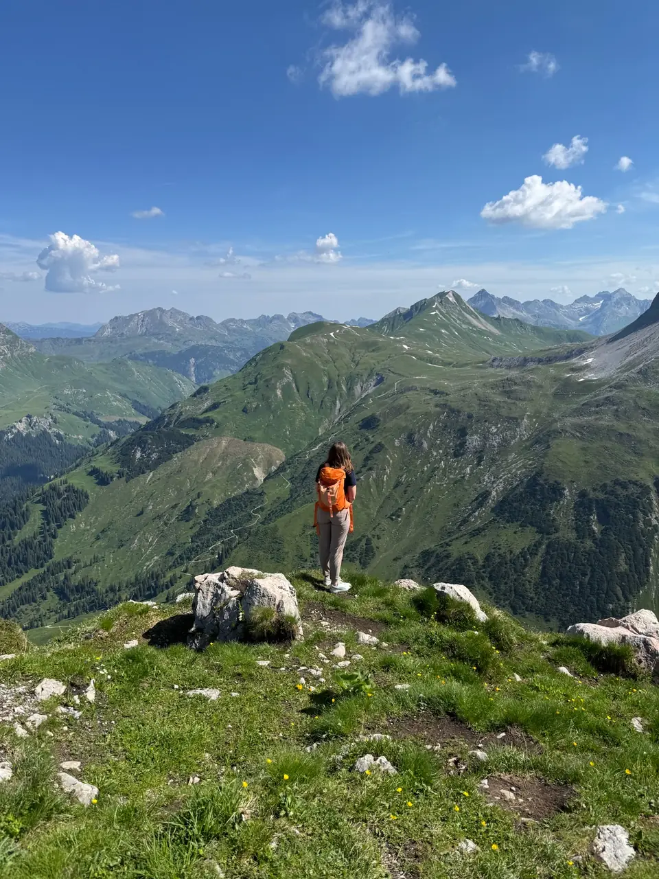 Wanderin mit orangefarbenem Tagesrucksack vor einem weiten Bergpanorama